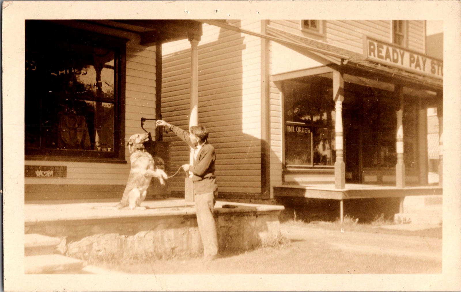 [62k] Postcard RPPC, Man Giving Dog Treat, Street Scene, Stores